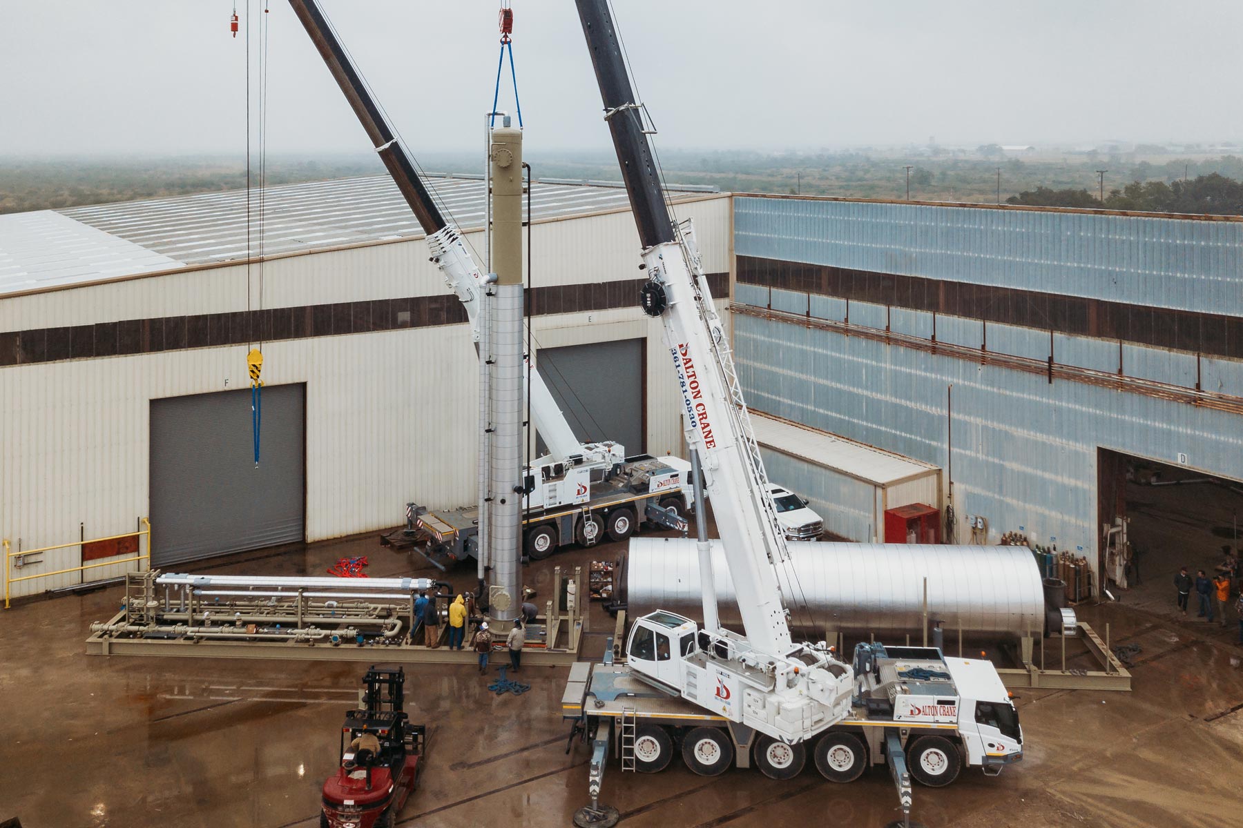 Crane lifting a large cylindrical structure outside an industrial warehouse on a cloudy day.